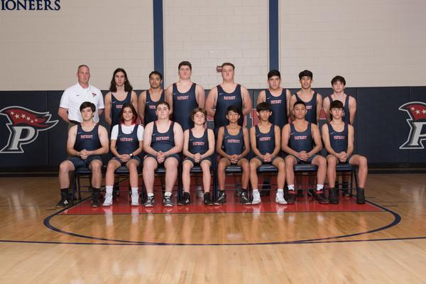 Two rows of junior varsity wrestlers dressed in blue singlets.  First row has eight, seated wrestlers.  Second row with seven standing wrestlers and coach in white polo shirt. 